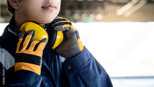 Asian man construction worker wearing uniform suit, safety helmet, goggles and protective gloves holding yellow ear muffs or ear defenders on his neck at construction site photo