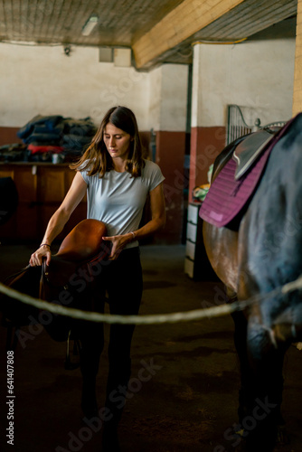 A rider puts a saddle on her black horse in the stables in preparation for the race equestrian concept  © Guys Who Shoot