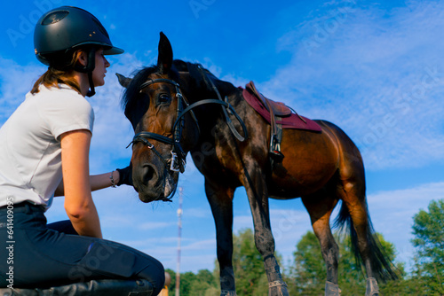 A horsewoman in a helmet hugs and scratches her beautiful black horse with her hand at the equestrian arena during a horseback ride