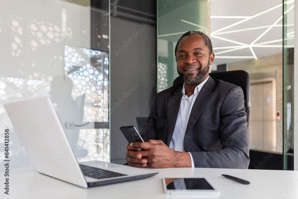 Portrait of senior gray haired african american boss, man smiling and looking at camera at workplace inside office, businessman holding phone, using online app on smartphone. Portrait of senior gray haired african american boss, man smiling and looking at camera at workplace inside office, businessman holding phone, using online app on smartphone.