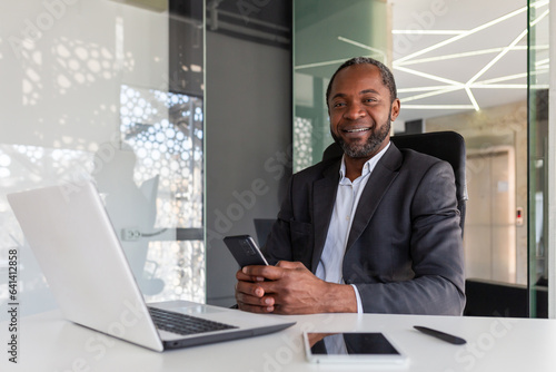 Portrait of senior gray haired african american boss, man smiling and looking at camera at workplace inside office, businessman holding phone, using online app on smartphone.