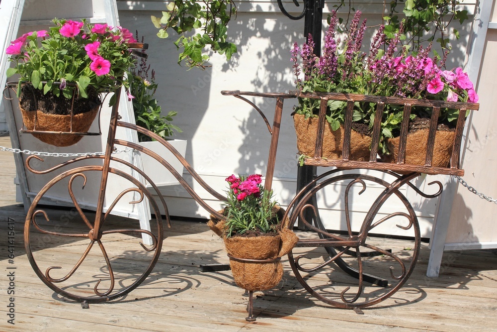 Summer decorative white bicycle with basket flowers in green garden. Flower pots in ornate bicycle in city park. Landscaping design. Decorative flower stand in the shape of a bicycle with flower pots