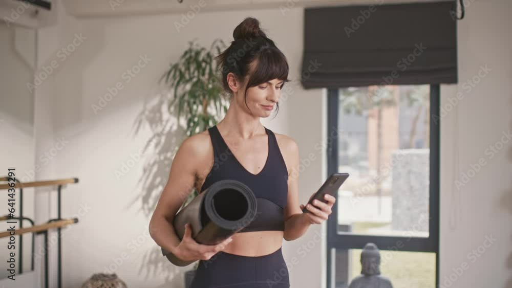 Beautiful Caucasian female fitness trainer chatting with her customer on phone while standing in sport center. Sporty woman holding mat during remote communication. Gym concept. Beautiful Caucasian female fitness trainer chatting with her customer on phone while standing in sport center. Sporty woman holding mat during remote communication. Gym concept.