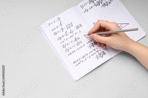 Woman writing maths formulas in copybook with pencil on grey background