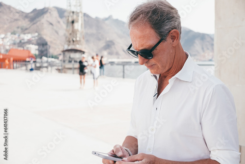 Handsome mature businessman in outdoors using smartphone typing a message. Smiling relaxed people in sunglasses and white shirt holding mobile phone enjoying tech and social #641437066