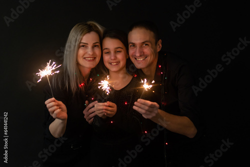 Happy family in black - woman, man and little girl, with a sparkler in hand the New Year\'s Christmas at home. The girl at the father on hands. New year 2023.