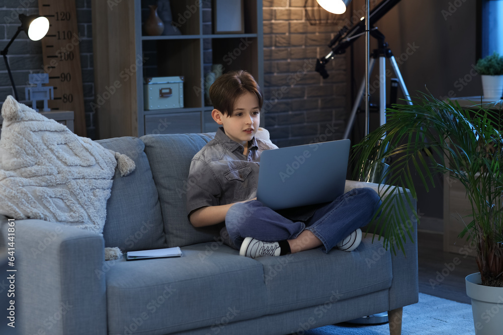 Little boy doing homework with laptop and sitting on sofa at home in evening Little boy doing homework with laptop and sitting on sofa at home in evening