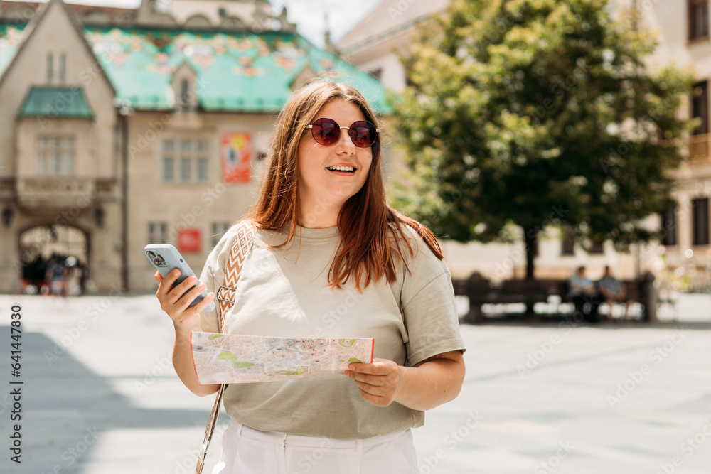 Happy tourist girl with map and smart phone travels in Europe. Vacation concept by exploring interesting places to travel. Redhead Woman Searching locations