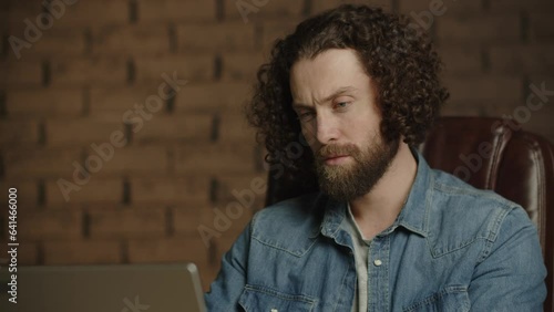 Close-up of a man working on a laptop. Begginer programmer write code, drink coffee, think. Work online in a spacious, large office. Confident curly styled freelancer drinking coffee at workplace photo