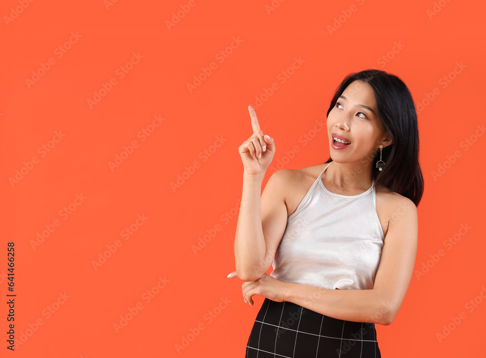Happy young beautiful Asian woman pointing at something on orange background Happy young beautiful Asian woman pointing at something on orange background