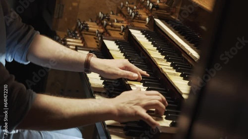 Expert organist plays a large church organ keyboard. Hands move quickly improvising sacred music. Musician presses keys and adjusts the sound with stops and rods and also uses foot pedals.