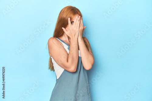 Redhead young woman on blue background blink through fingers frightened and nervous.