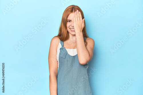 Redhead young woman on blue background having fun covering half of face with palm.