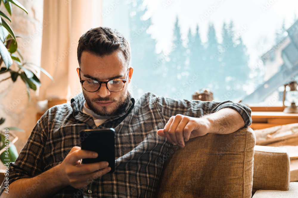 Young adult using mobile phone while sitting indoors. Focused man typing on digital device feeling hesitant about his decisions. Young adult using mobile phone while sitting indoors. Focused man typing on digital device feeling hesitant about his decisions.