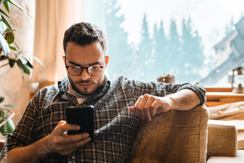 Young adult using mobile phone while sitting indoors. Focused man typing on digital device feeling hesitant about his decisions.