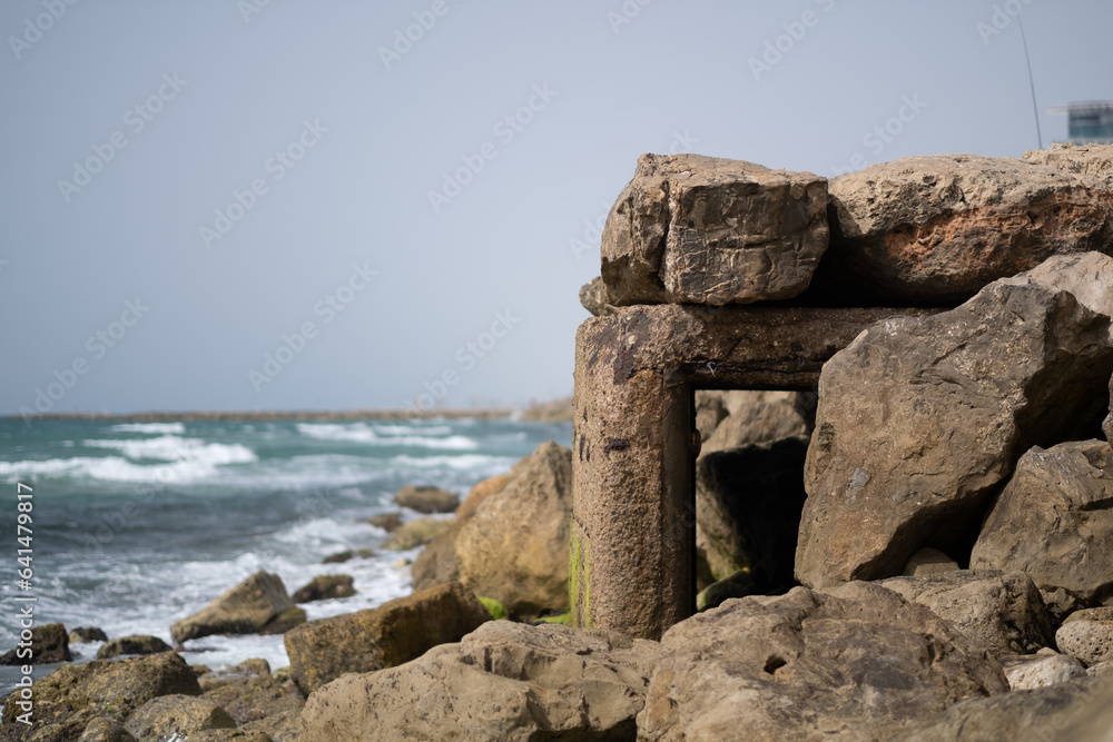 A concrete cube amid the boulders, creating a retaining wall for the boardwalk in Tel Aviv. In the distance, waves are crashing at the shore of the Mediterranean Sea. A concrete cube amid the boulders, creating a retaining wall for the boardwalk in Tel Aviv. In the distance, waves are crashing at the shore of the Mediterranean Sea.