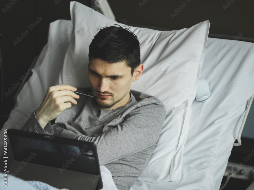 A young man with a tablet lies in bed in a hospital.