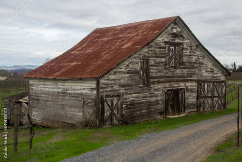 Wallpaper Mural An old weather worn wooden barn with a red metal roof stands beside a dirt road. Green grass is around the side of the barn. A cloudy sky is behind the barn Torontodigital.ca