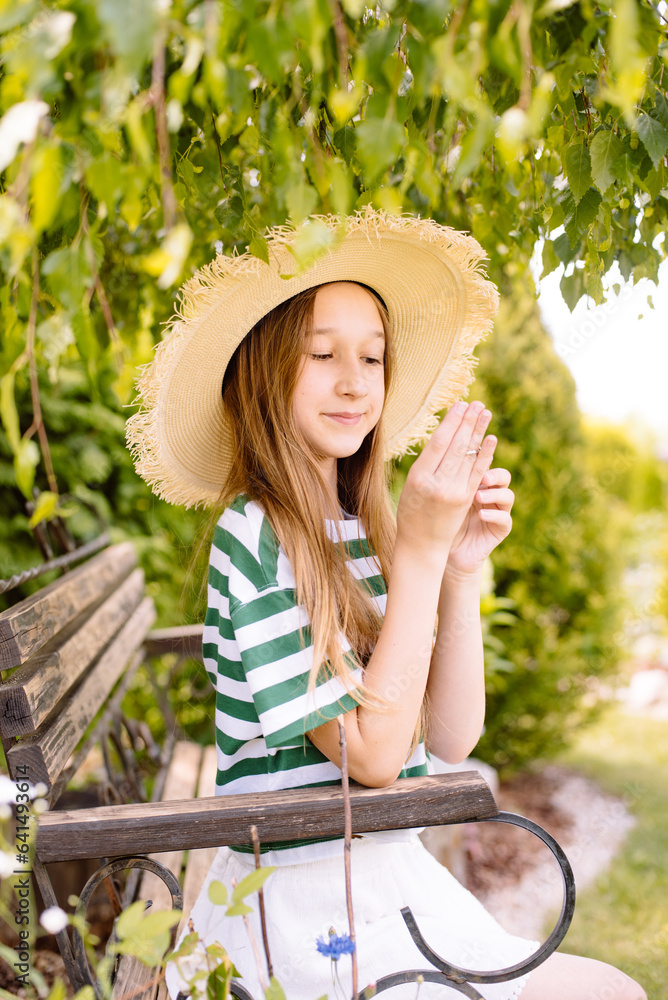 Obraz premium A girl in a hat with long hair is sitting on a bench in a green garden and resting after work