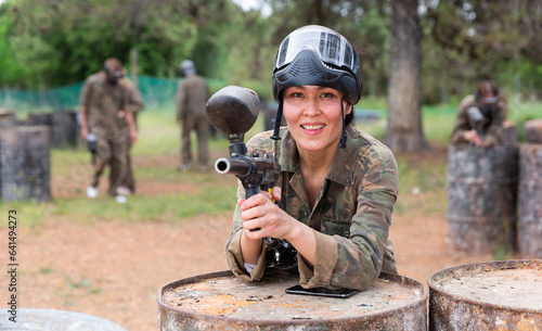 Smiling woman wearing uniform and holding gun ready for playing paintball with friends outdoor