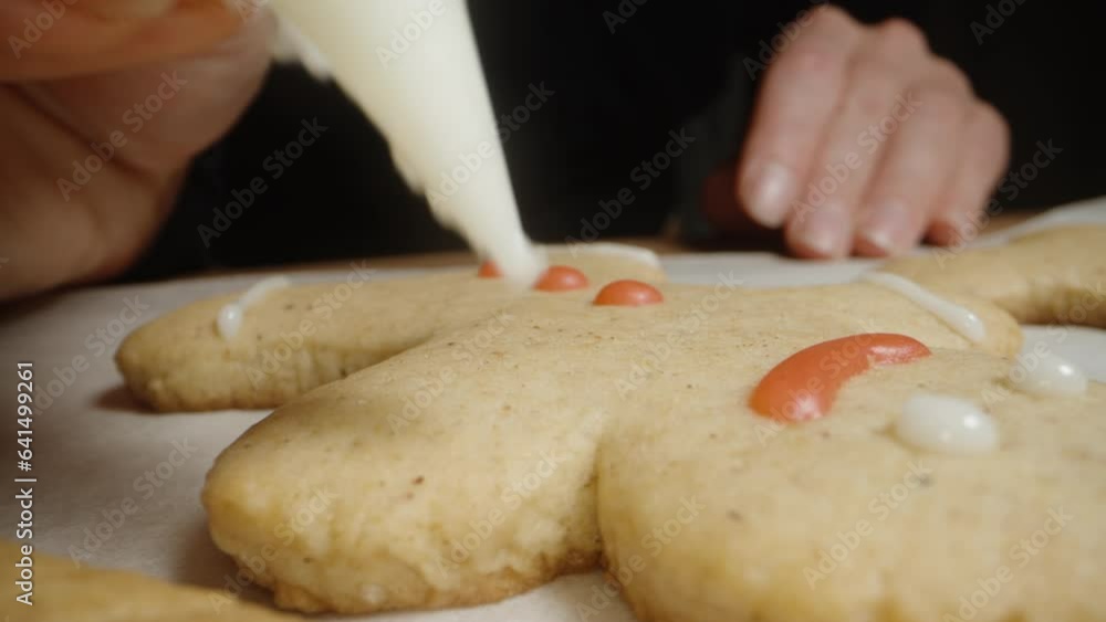 From a pastry bag, the woman squeezes a thin stream of white icing onto gingerbread cookies shaped like little people. Close-up view. From a pastry bag, the woman squeezes a thin stream of white icing onto gingerbread cookies shaped like little people. Close-up view.