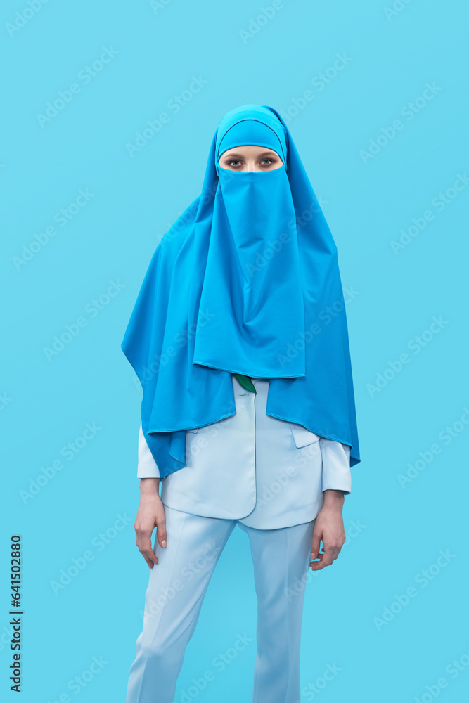 Portrait of unrecognizable woman in elegant suit and blue niqab standing against blue background and looking at camera Portrait of unrecognizable woman in elegant suit and blue niqab standing against blue background and looking at camera