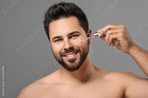 Handsome man applying cosmetic serum onto face on grey background