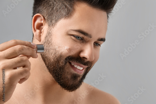 Handsome man applying cosmetic serum onto face on grey background, closeup