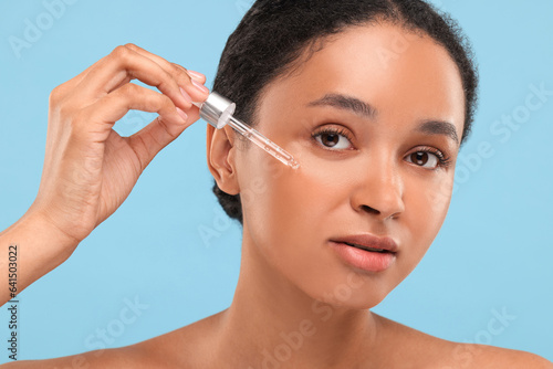 Beautiful woman applying serum onto her face on light blue background, closeup