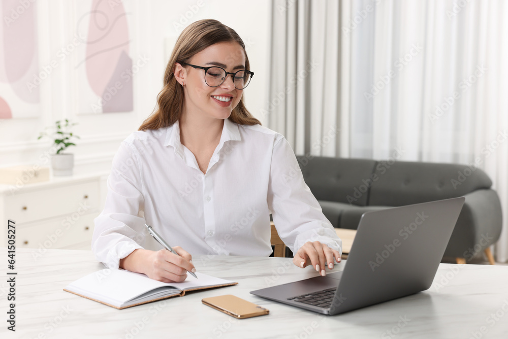 Happy woman with notebook and laptop at white table in room Happy woman with notebook and laptop at white table in room