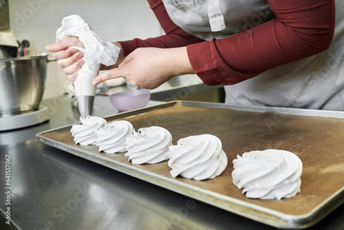 Unrecognizable female pastry chef using piping bag while making meringue nests in confectionery kitchen 
