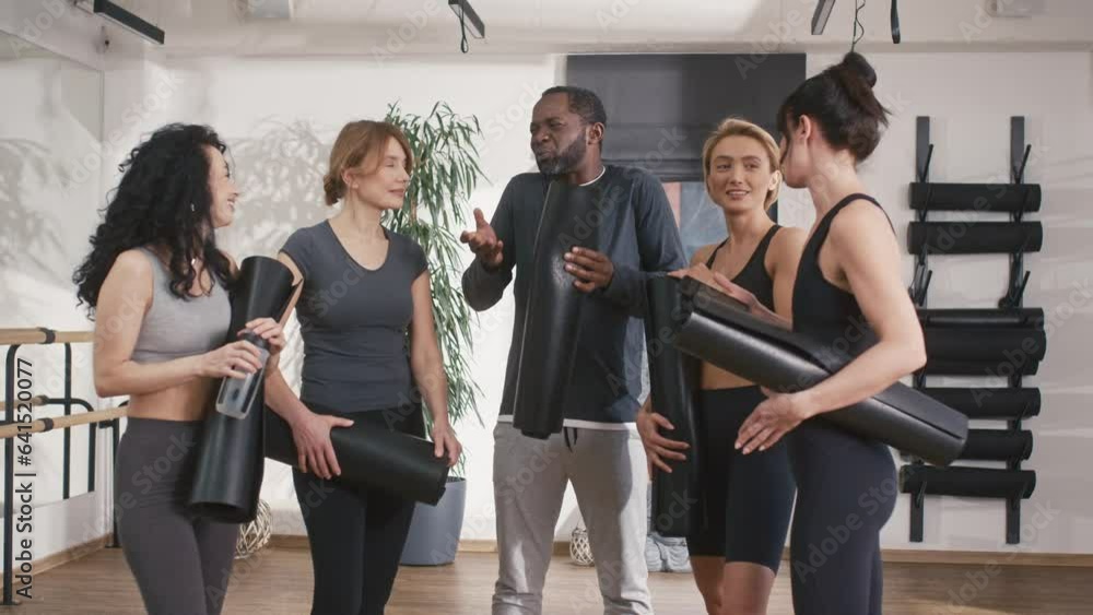 Positive mixed-race people with fitness suits communicating with each other in pilates studio. Having funny conversation before training. Friends speaking together while holding sports mats. Positive mixed-race people with fitness suits communicating with each other in pilates studio. Having funny conversation before training. Friends speaking together while holding sports mats.