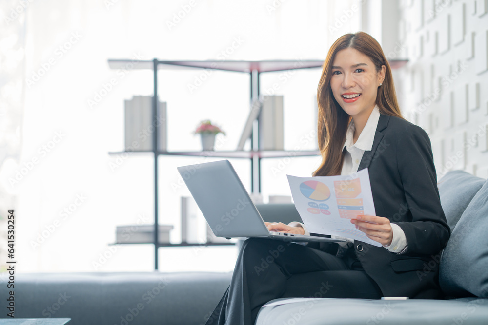 Image of a young beautiful Asian Business woman smiling while working with a laptop and talking to work doing math finance on an office desk, tax, report, accounting, statistics, and research concepts Image of a young beautiful Asian Business woman smiling while working with a laptop and talking to work doing math finance on an office desk, tax, report, accounting, statistics, and research concepts