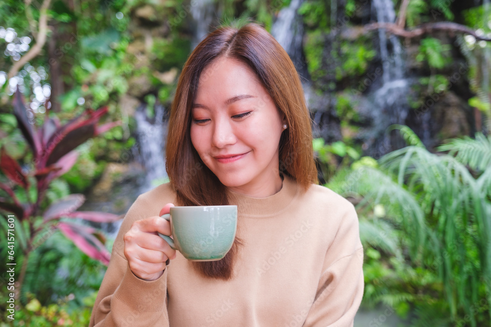 Portrait image of a young woman holding and drinking coffee while sitting in the garden with waterfall Portrait image of a young woman holding and drinking coffee while sitting in the garden with waterfall