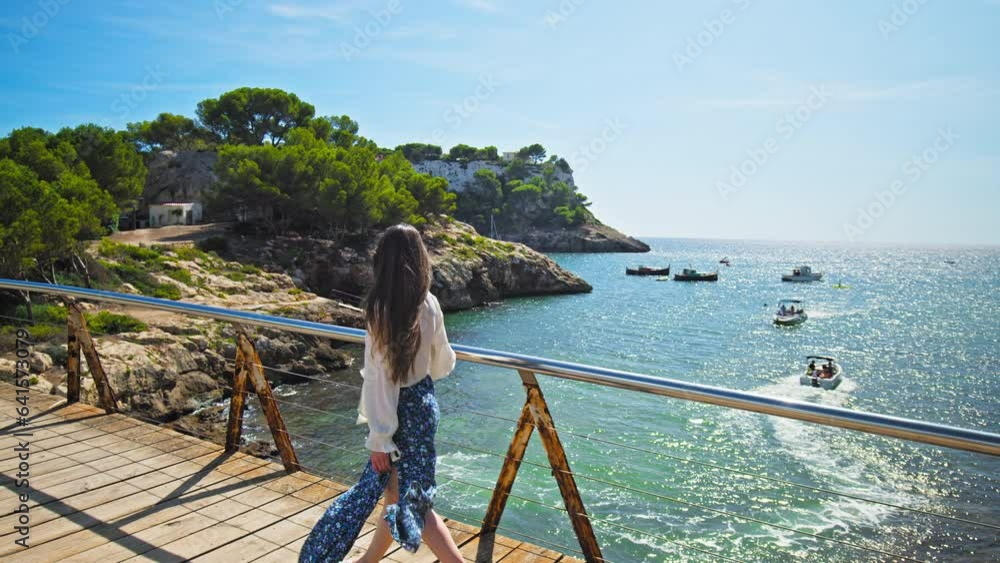 Beautiful girl walking on a wooden bridge in Cala Galdana overlooking the beach. Wind blowing a young woman with long hair and dress, relaxing luxury lifestyle concept on the Spanish Balearic Islands. Beautiful girl walking on a wooden bridge in Cala Galdana overlooking the beach. Wind blowing a young woman with long hair and dress, relaxing luxury lifestyle concept on the Spanish Balearic Islands.