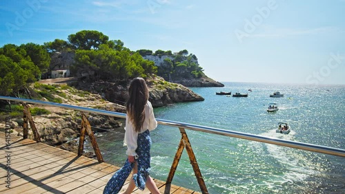 Beautiful girl walking on a wooden bridge in Cala Galdana overlooking the beach. Wind blowing a young woman with long hair and dress, relaxing luxury lifestyle concept on the Spanish Balearic Islands. photo
