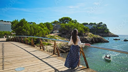 Beautiful girl walking on a wooden bridge in Cala Galdana overlooking the beach. Wind blowing a young woman with long hair and dress, relaxing luxury lifestyle concept on the Spanish Balearic Islands. photo