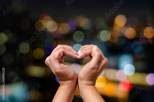Female hand making heart symbol on night city colorful bokeh background. Love concept on Valentine day.