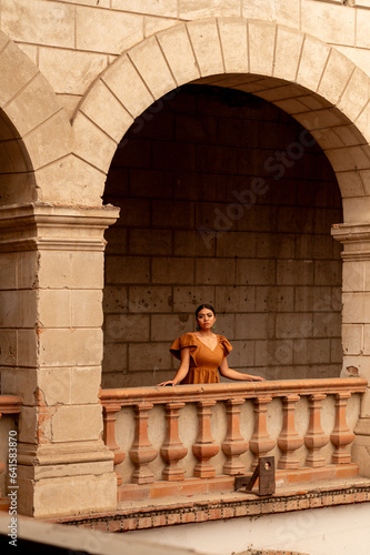 Frida-Inspired Essence: A Mexican woman, reminiscent of Frida Kahlo, graces an aged chair beneath delicate archways. Bathed in a play of light and shadow, her traditional attire and contemplative gaze