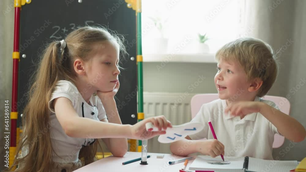 Caring long haired girl fans little brother with number cards in classroom on hot day. Sister helps toddler kid to cool down at homework in nursery Caring long haired girl fans little brother with number cards in classroom on hot day. Sister helps toddler kid to cool down at homework in nursery