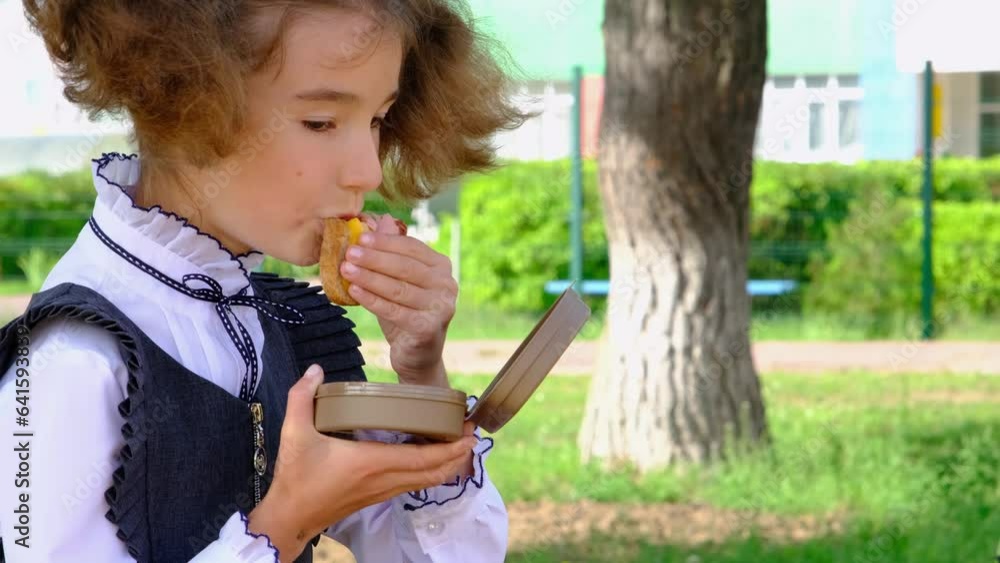 Girl with backpack eating sandwich packed in a sandwich box near school. A quick snack with a bun, unhealthy food, lunch from school. Back to school. Education, primary school classes, September 1 Girl with backpack eating sandwich packed in a sandwich box near school. A quick snack with a bun, unhealthy food, lunch from school. Back to school. Education, primary school classes, September 1