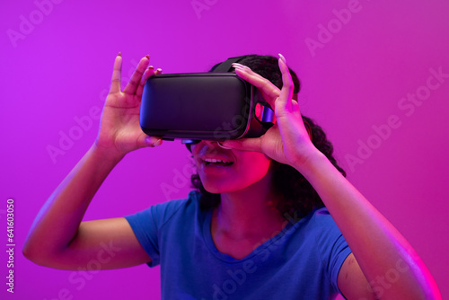 Biracial woman using vr headset on neon pink to purple background