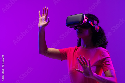 Biracial woman using vr headset and pointing on neon pink to purple background © vectorfusionart