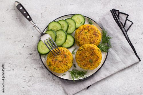Delicious millet cutlets with carrots and seeds served with cucumber and herbs on a gray textured background, top view. Homemade vegan food