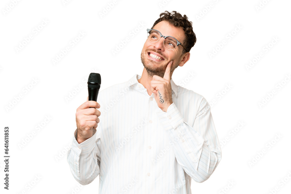 Brazilian man picking up a microphone over isolated chroma key background thinking an idea while looking up Brazilian man picking up a microphone over isolated chroma key background thinking an idea while looking up