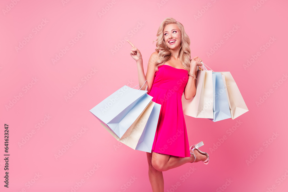Photo of cute lovely funny girl hold mall shop bags look indicate finger empty space wear mini dress isolated on pink color background