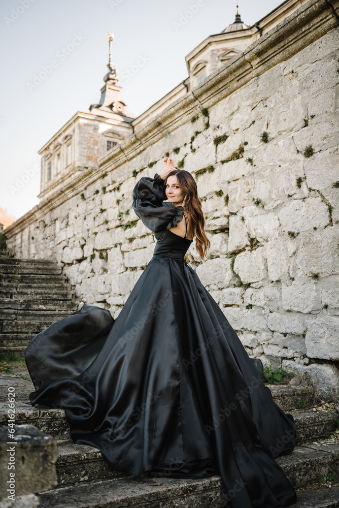 Female in long black dress stands on stairs near ancient palace at sunset. Model girl in nature autumn day. Luxury woman near old Pidhirtsi Castle, Lviv region, Ukraine. Stylish bride. Back view