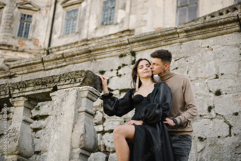 Woman in black long dress hugs man stand on stairs near ancient palace. Female kissing male in street. Luxury couple newlyweds near old Pidhirtsi Castle, Lviv, Ukraine. Stylish bride and groom outdoor Woman in black long dress hugs man stand on stairs near ancient palace. Female kissing male in street. Luxury couple newlyweds near old Pidhirtsi Castle, Lviv, Ukraine. Stylish bride and groom outdoor