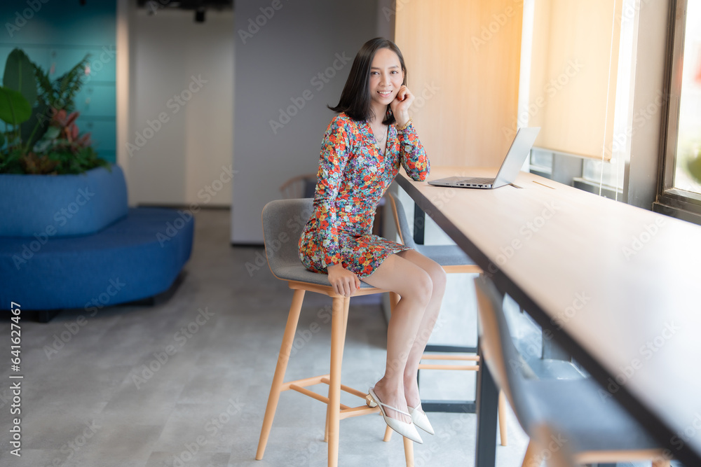 Asian female executive with short hair, company boss wearing a floral suit, sending a beautiful smile, having a laptop on the desk for business Sitting in a chair near the window in the office space Asian female executive with short hair, company boss wearing a floral suit, sending a beautiful smile, having a laptop on the desk for business Sitting in a chair near the window in the office space