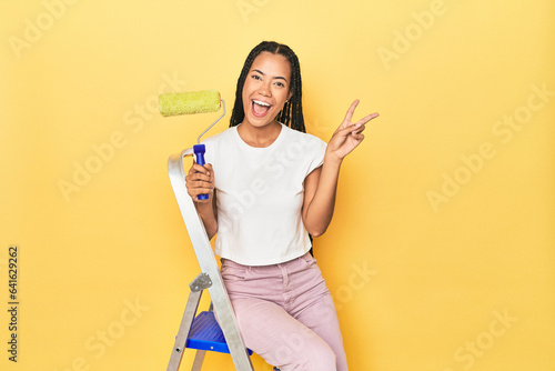 Indonesian woman with roller on ladder on yellow joyful and carefree showing a peace symbol with fingers.